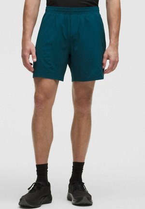 Mannelijke benen die donker teal sportshorts, zwarte enkelsokken en zwarte hardloopschoenen dragen, staand tegen een effen lichte achtergrond.