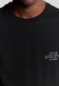 Sort bomulds T-shirt med klassisk rund halsudskæring. Har hvidt trykt tekst på venstre bryst, der læser "LYLE ATHLETIC FUEL AMBITION."