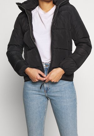 Person trägt eine schwarze Daunenjacke über einem weißen T-Shirt und hellblauen Jeans, schließt den Reißverschluss der Jacke an der Taille.