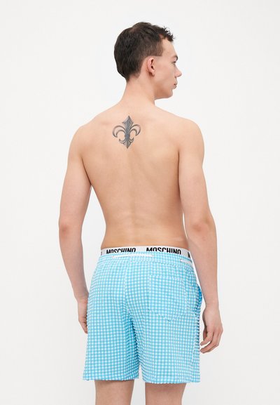 Zilā gingham peldbikses ar melnu Moschino jostu. Iespējojams aizmugures kabata un četrkantainais raksts. Modelis ir bez krekla, skatīts no aizmugures.