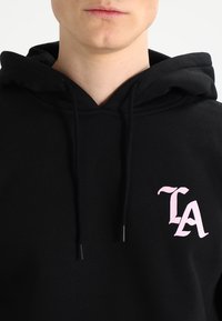 Zwarte hoodie met een trekkoord, voorzien van een roze "TA" logo aan de linkerkant. Het materiaal lijkt zacht en textuurrijk.