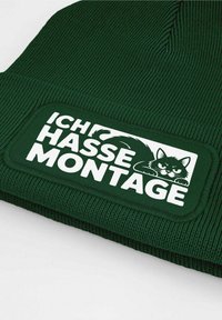 Green beanie with ribbed texture featuring a white rectangular patch. The patch displays the text "ICHI HASSE MONTAGE" and an illustration of a cat.