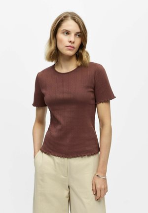 Femme aux cheveux blonds portant un t-shirt marron ajusté à manches courtes et un pantalon beige, debout, les mains dans les poches, devant un fond blanc.