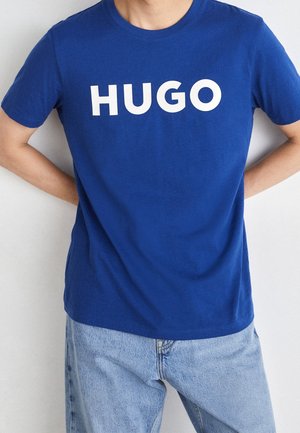 Osoba nosząca niebieski T-shirt z "HUGO" białymi literami i jasnoniebieskie dżinsy, stojąca z rękami za plecami.