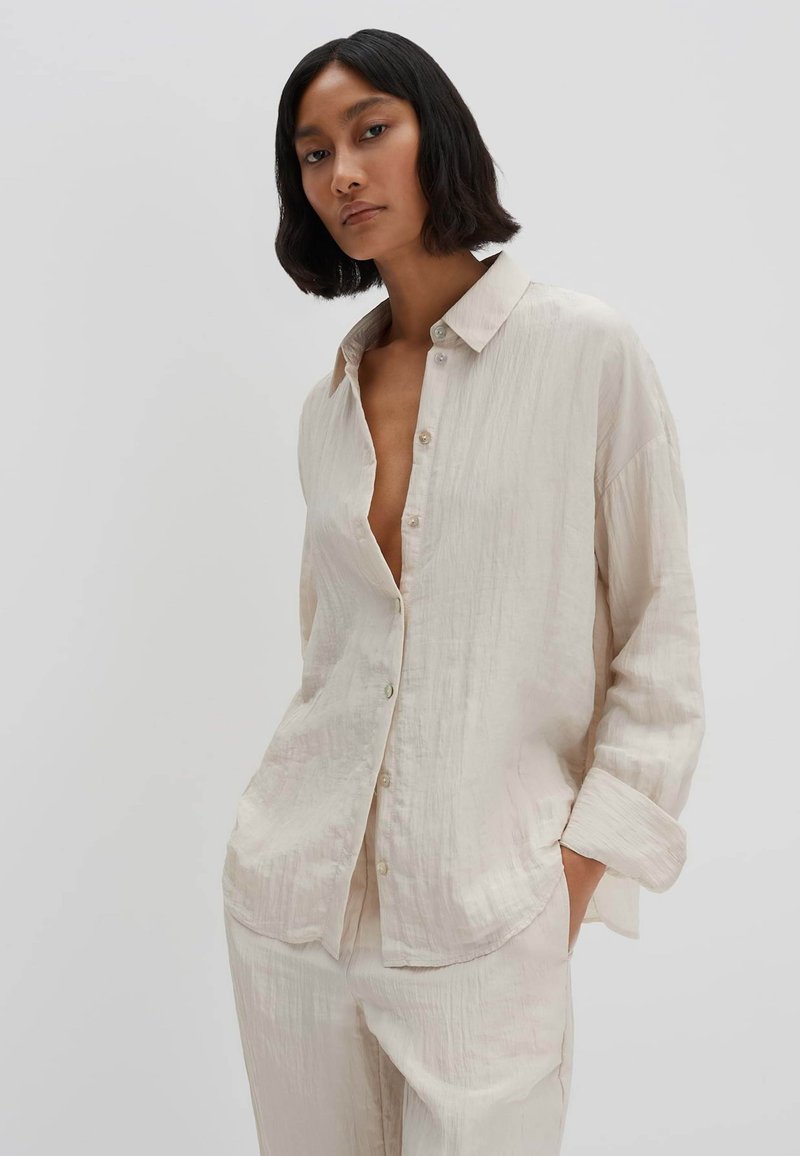 Camicia button-up oversize beige chiaro in un tessuto texture. Presenta un colletto e tasche sul petto, abbinata a pantaloni coordinati.