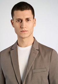 Blazer de color marrón claro con tejido texturizado y solapas con muesca, combinado con una camiseta blanca. Presenta un bolsillo lateral y líneas limpias.