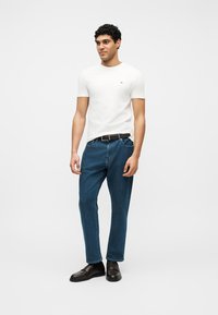 Νεαρός άνδρας που στέκεται, φοράει λευκό μπλουζάκι, μπλε τζιν με μαύρη ζώνη και σκούρα καφέ παπούτσια, σε απλό λευκό φόντο.