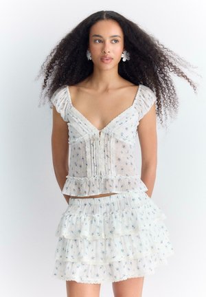 Femme aux longs cheveux bouclés portant un haut blanc à volants fleuris et une jupe mini à volants assortie, debout devant un fond clair uni.