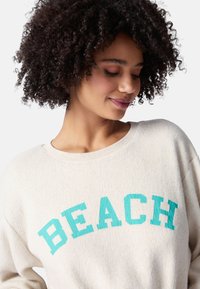 Sudadera de color crema con una textura suave y un ajuste relajado. Presenta un texto en negrita de color teal que dice "BEACH" en la parte frontal.