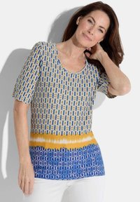 Korte mouwen, gebloemd top met blauwe, witte en gele kleuren; heeft een ronde halslijn en verschillende texturen in geometrische ontwerpen.