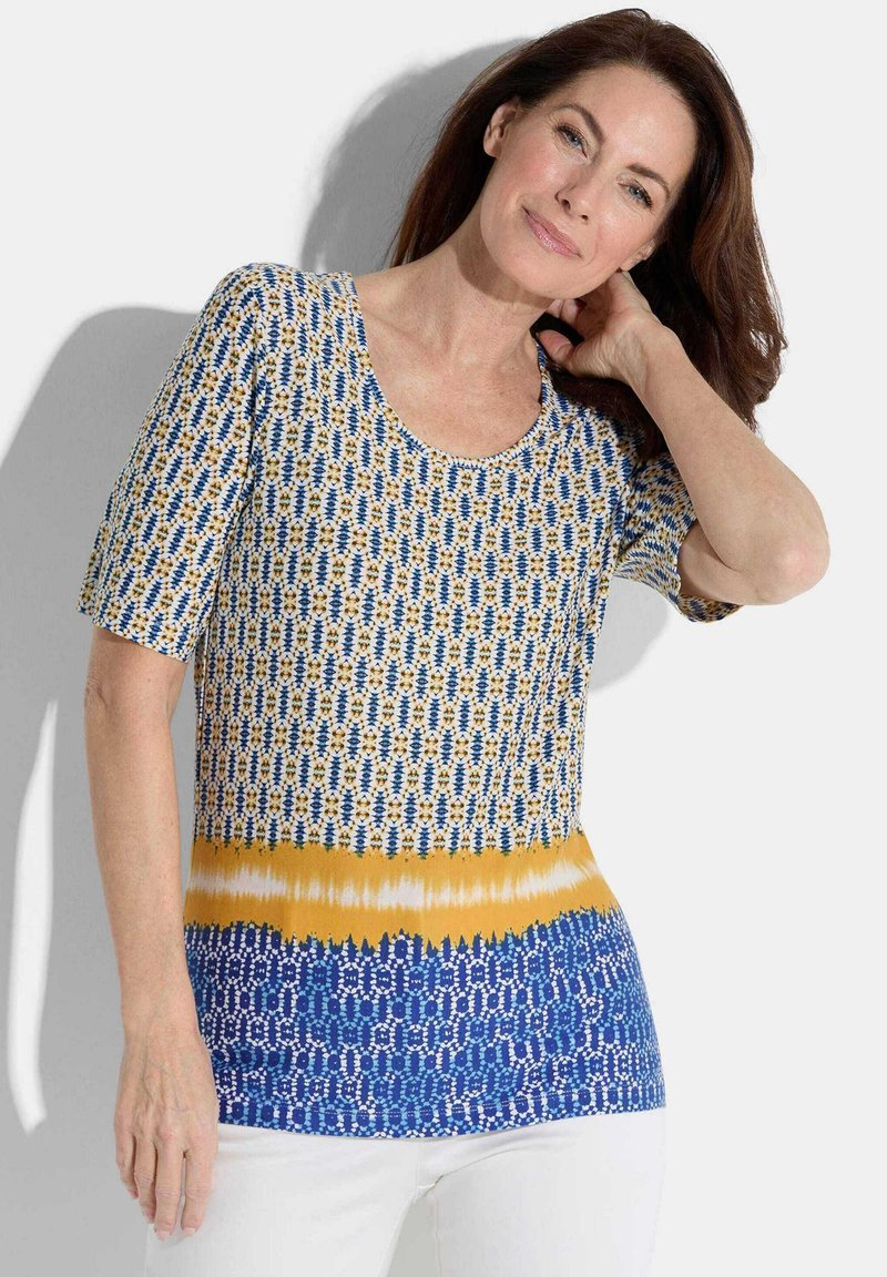 Korte mouwen, gebloemd top met blauwe, witte en gele kleuren; heeft een ronde halslijn en verschillende texturen in geometrische ontwerpen.