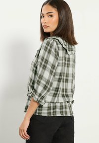Joven con cabello castaño hasta los hombros, vistiendo una blusa de cuadros verde y blanca con cuello con volantes y pantalones negros, mirando de lado.