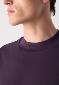 Artimas žmogaus, dėvinčio violetinę marškinių su apvaliu kaklu, kuriame centre yra mažas juodas "BOSS" logotipas.