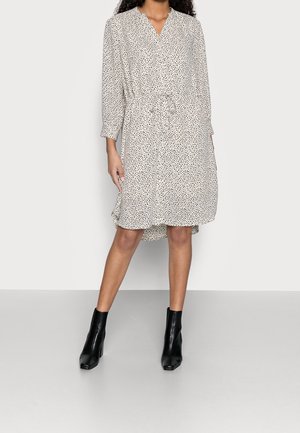 Femme portant une robe blanche mi-genou avec de petits pois noirs, taille cintrée, et des bottines noires, debout devant un fond uni.