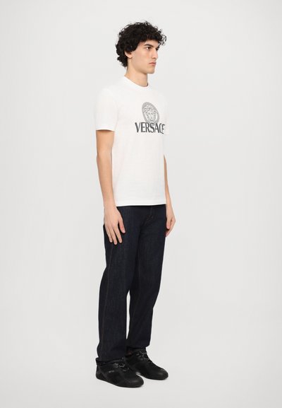 Hvid bomulds t-shirt med korte ærmer, med et sort trykt Medusa-logo og "VERSACE" tekst. Bæres med mørkeblå jeans og sorte sko.