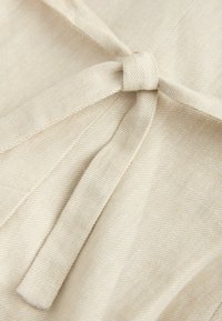 Primer plano de tela beige con un nudo simple atado en una tira estrecha, mostrando textura y detalles de costura diagonal.