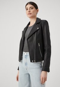 Svart skinnbikerjakke med en asymmetrisk glidelås, sølvfarget hardware og skreddersydd passform, kombinert med en grå topp og lyseblå jeans.