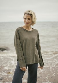 Suéter de punto verde oliva con cuello redondo y mangas largas, que presenta seis grandes botones decorativos en los puños. Combinado con jeans oscuros.