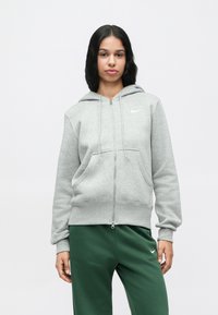 Sieviete, tērpusies gaiši pelēkā ar rāvējslēdzi, un tumši zaļās biksēs, stāv pretī vienkāršai baltai fona.