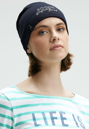 Young woman with short brown hair wears a navy beanie with white text "SOCCX Original Brand" and a striped shirt with partially visible text.