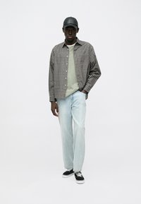 Camicia a quadri nei toni del marrone e crema sopra un maglione verde chiaro, abbinata a jeans azzurri e sneakers nere con suole bianche.