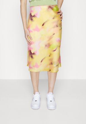 Frau trägt einen gelben Midi-Rock mit floralem Muster in Pink, Grün und Braun, kombiniert mit weißen Turnschuhen, stehend vor einem schlichten Hintergrund.