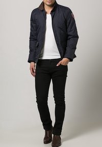 Marineblau gesteppte Jacke mit braunem Kragen, Knopfverschluss vorne, Seitentaschen und Stoff mit Diamantmuster. Getragen über einem weißen T-Shirt und schwarzen Jeans.