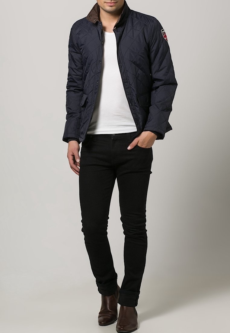 Marineblau gesteppte Jacke mit braunem Kragen, Knopfverschluss vorne, Seitentaschen und Stoff mit Diamantmuster. Getragen über einem weißen T-Shirt und schwarzen Jeans.