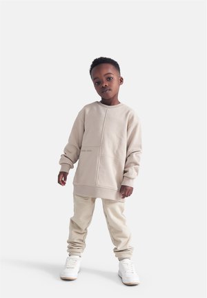 Dreng stående med afslappede hænder, iført beige oversized sweatshirt, matchende bukser og hvide sneakers på en ensfarvet lys baggrund.