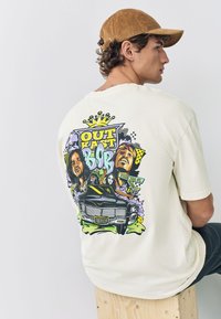 Kermanvärinen T-paita, jonka takana on värikäs kuva kahdesta hahmosta ja autosta. Lyhyet hihat ja rento istuvuus.