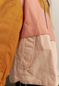 Färgglad jacka med orange och rosa paneler; texturerad tyg med rutmönster; har en ficka och en rund knappdetalj.