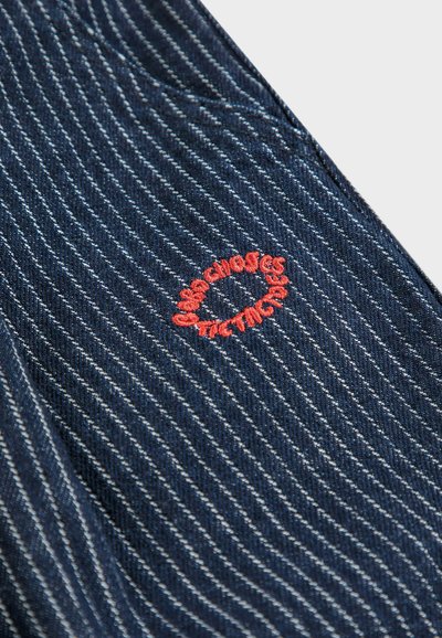 Denimtyg i mörkblått med vita ränder; har en cirkulär röd broderad logotyp med texten "chaos" och "rules."