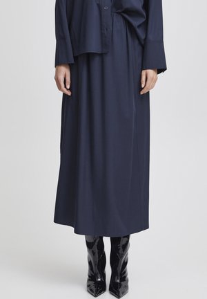 Kobieta nosi granatową maxi spódnicę i dopasowaną bluzkę z długim rękawem oraz czarne lakierowane botki z ostro zakończonym noskiem na białym tle.