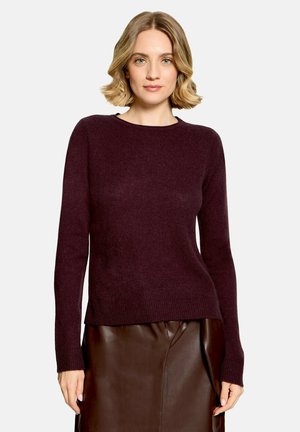 Femme aux cheveux blonds mi-longs portant un pull violet foncé à manches longues et une jupe en cuir marron, debout devant un fond clair.