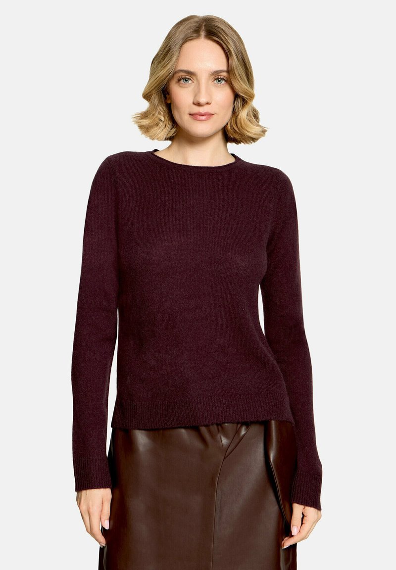 Femme aux cheveux blonds mi-longs portant un pull violet foncé à manches longues et une jupe en cuir marron, debout devant un fond clair.
