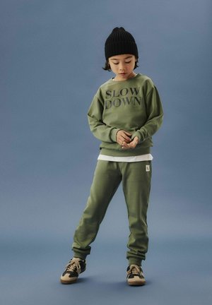Enfant portant un sweat-shirt olive avec l'inscription "Ralentir", un pantalon de jogging assorti, un bonnet noir et des baskets, se tenant devant un fond bleu uni.