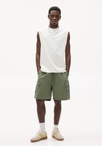 Chemise sans manches blanche, shorts cargo vert olive avec poches latérales, chaussettes blanches, baskets beiges avec accents jaunes. Fond neutre.