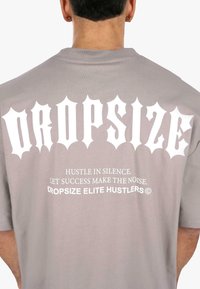 Grå kortærmet t-shirt med stort hvidt gotisk tekst på ryggen, med en glat bomuldsstruktur og en afslappet pasform.