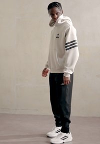 Lys beige hoodie med sort tre-stribet detalje, sorte bukser og hvide atletiske sko. Materialet ser blødt ud; designet er afslappet og tætsiddende.