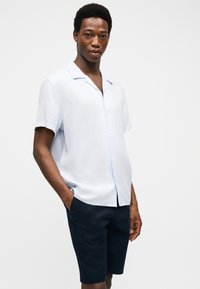 Bărbat tânăr stând cu mâinile în buzunare, purtând o cămașă albastru deschis cu mânecă scurtă și pantaloni scurți albastru închis, pe un fundal alb.