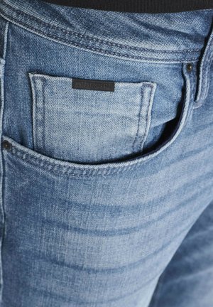 Svijetloplave traperice s velikim stražnjim džepom i vidljivom crnom etiketom. Suptilna izbljeđena područja i teksturne detalje poboljšavaju izgled tkanine.