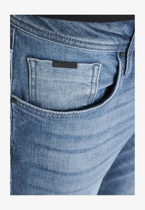 Jeans in denim di un blu chiaro con una grande tasca posteriore e un'etichetta nera visibile. Leggeri effetti di sbiadimento e dettagli di texture arricchiscono l'aspetto del tessuto.