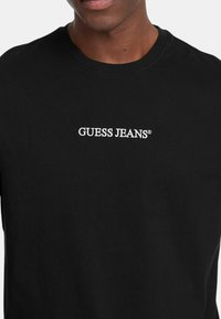 T-shirt in cotone nero con logo ricamato bianco "GUESS JEANS®" sul petto, scollatura tonda e maniche corte.