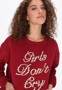 Bordó pulóver gyöngyözött "Girls Don't Cry" felirattal, bordázott kerek nyakkal és puha tapintással. Göndör haj és fülbevalók láthatók.