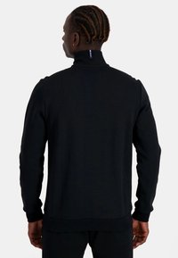 Zwarte hoodie met rits, voorzien van een hoge kraag en geribde manchetten. De stof lijkt glad, met een klein logotag aan de achterkant van de halslijn.