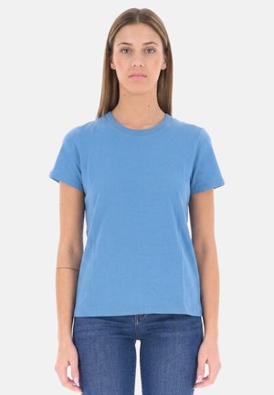 Donna in piedi rivolta in avanti, che indossa una t-shirt blu tinta unita a maniche corte e jeans blu scuro su sfondo bianco.