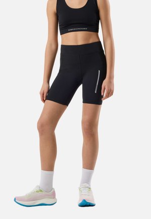Femme portant un soutien-gorge de sport noir Swedemount et un short de cyclisme noir avec une bande réfléchissante, des chaussettes blanches et des chaussures de course roses.