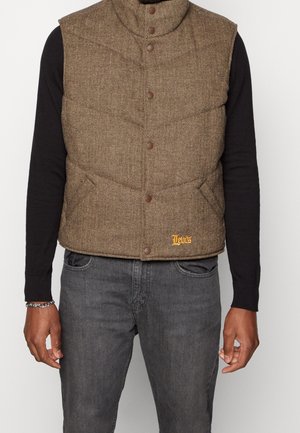 Veste matelassée marron en chevron avec boutons-pression portée sur une chemise noire à manches longues et associée à un jean gris foncé.