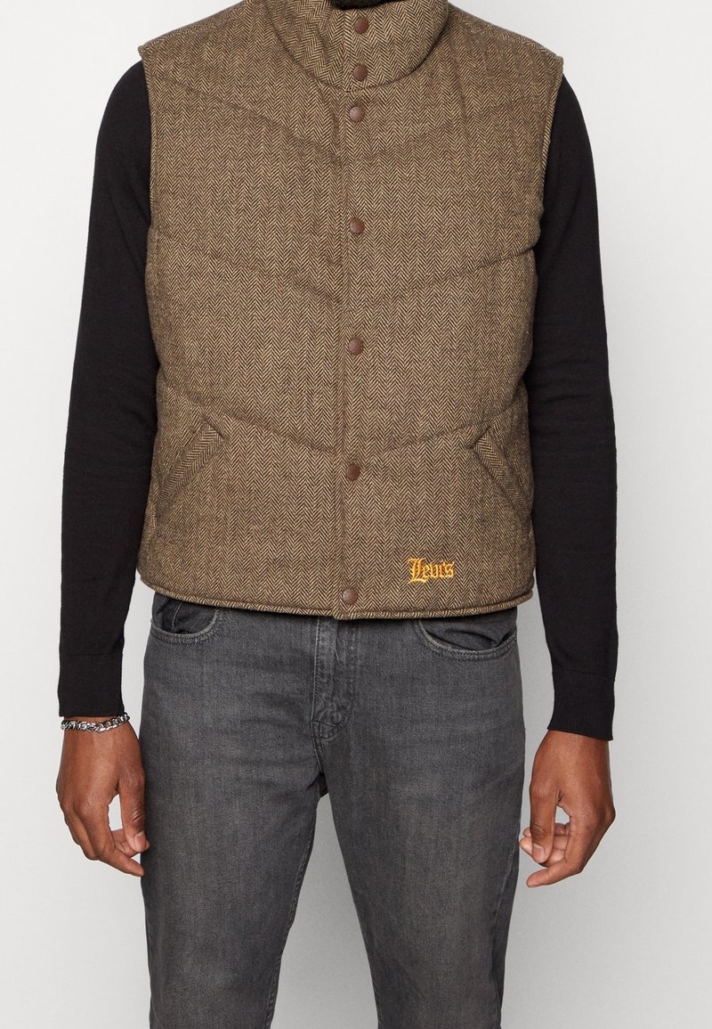 Veste matelassée marron en chevron avec boutons-pression portée sur une chemise noire à manches longues et associée à un jean gris foncé.