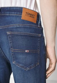 Mörkblå jeans i denim med en brun läderlapp märkt "TOMMY JEANS" på baksidan. Har synliga sömmar och en liten märkt etikett på fickan.